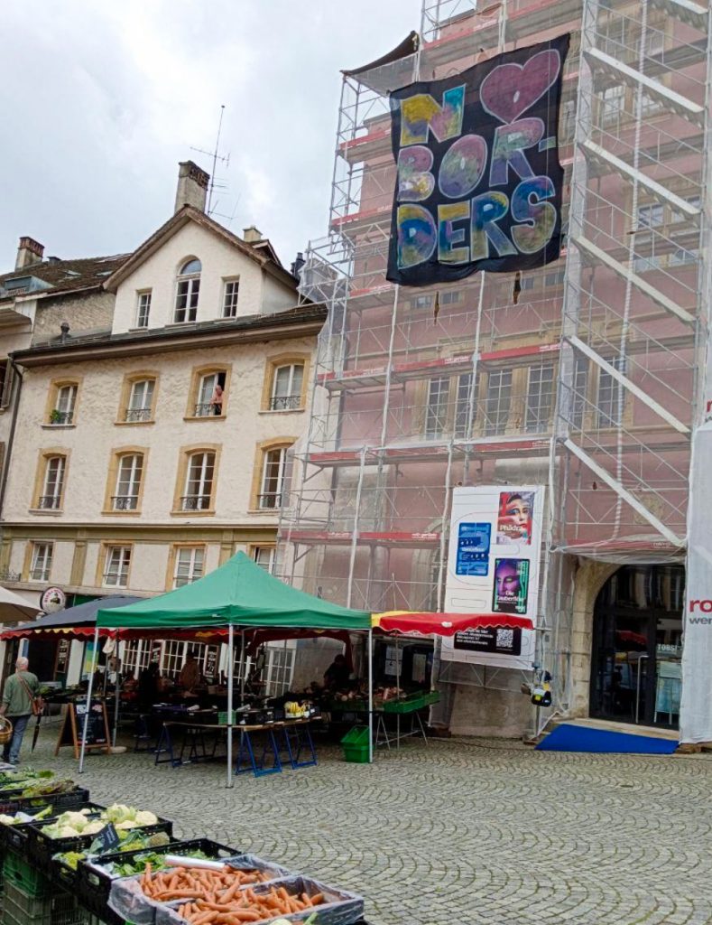 Ein grosses Banner hängt an einem Baugerüst mit der Aufschrift «No Borders» wobei das «o» von «No» eine Herz-Form hat. Unten auf der Strasse stehen Marktstände