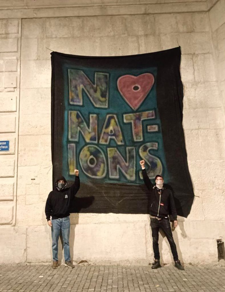 Zwei vermumte Personen stehen mit erhobener Faust vor einem Banner mit der Aufrsichft «No Nations», wobei das «o» von «No» ein Herz ist. Es ist Nacht.