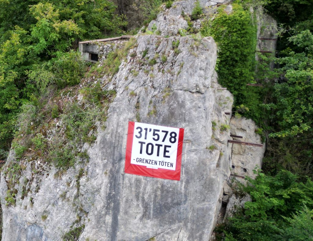 Eine grosse Schweizer Fahne befestigt an einer Felswand, fast vollständig überdeckt mit dem Schriftzug «31’578 Tote – Grenzen töten»