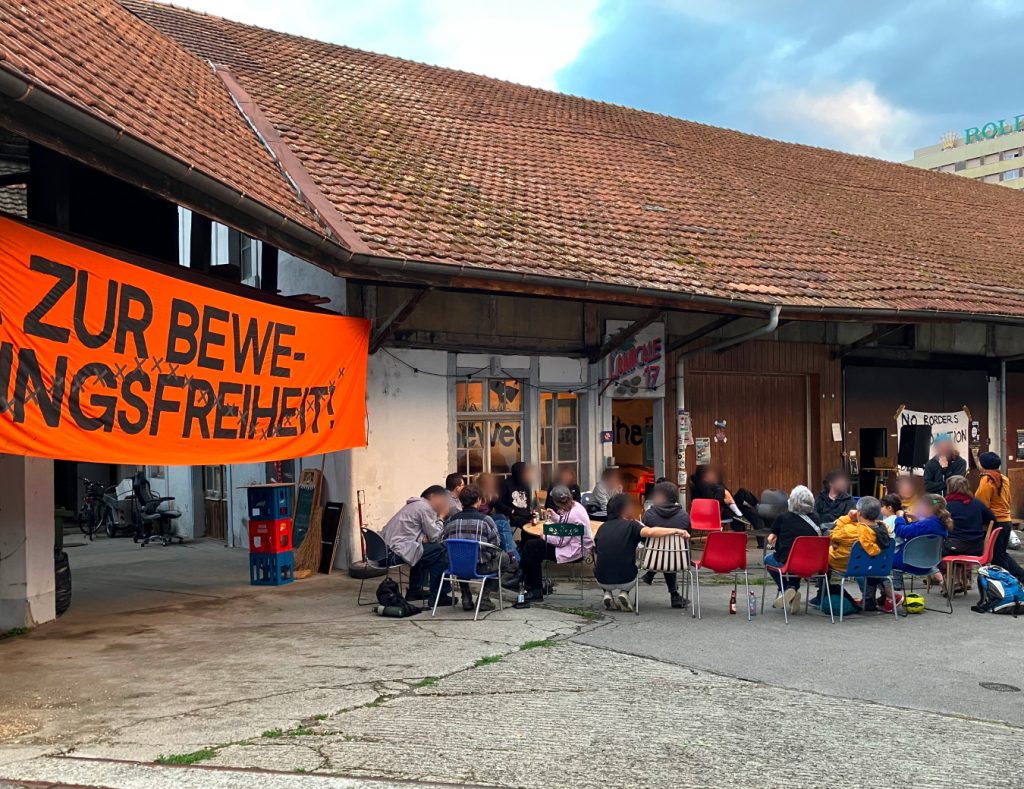 Vor dem Amicale im Quai du Bas sitzen Menschen auf Stühlen in einem Kreis, in verschieden Gespräche vertieft. Auf der Seite hängt ein Transparent mit der Aufschrift «Ja zur Bewegungsfreiheit»