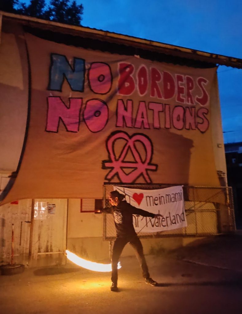 Eine Person schwingt während einer Feuershow ein brennender Ballen an einer Schnur, im Hintergrund ein grosses Transparent mit der Aufschrift «No Borders No Nations». Es ist Dämmerung, die Szene ist erhellt vom Feuer.