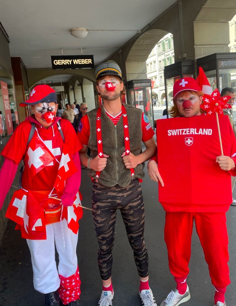 Drei Clowns in von Kopf bis Fuss eingekleidet in Schweizer Farben, Fähnchen, Wappen usw. posieren vor der Kamera. Der Clown in der Mitte trägt zusätzlich Armee-Camouflage.