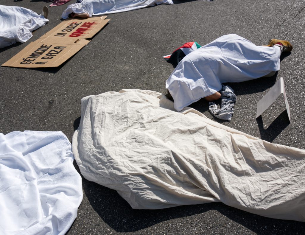 Körper unter weissen Tüchern liegen auf dem Asphalt, im Hintergrund ein Kartonschild mit der Aufschrift «Génocide à Gaza, La Suisse Complice»
