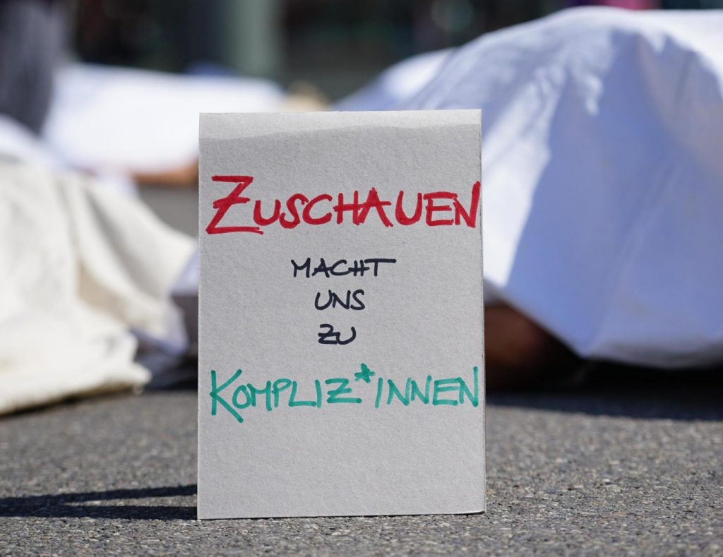 Schild steht auf dem Asphalt mit der Aufschrift «Zuschauen macht un Kompliz*innen». Im Hintergrund Körper unter weissen Tüchern